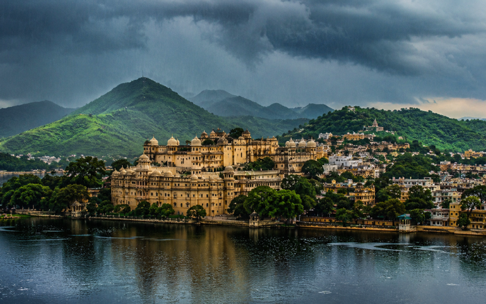 View of Udaipur City Palace and Lake Pichola surrounded by green Aravalli Hills under dramatic monsoon clouds in Rajasthan.