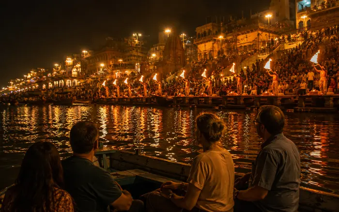 Tourists sitting in a wooden boat on the Ganges River watching the Ganga Aarti ceremony at Varanasi ghats, with fire lamps and illuminated temples reflecting on the dark water