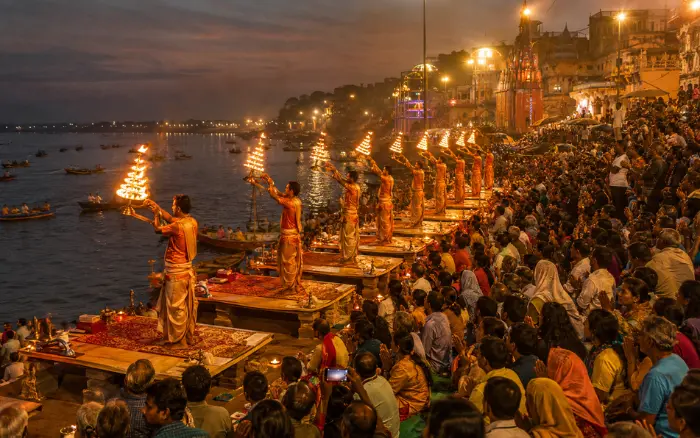 Seven priests in saffron robes performing the synchronized Ganga Aarti fire ceremony at Dashashwamedh Ghat in Varanasi, with large crowds watching from the stone steps beside the Ganges River at dusk
