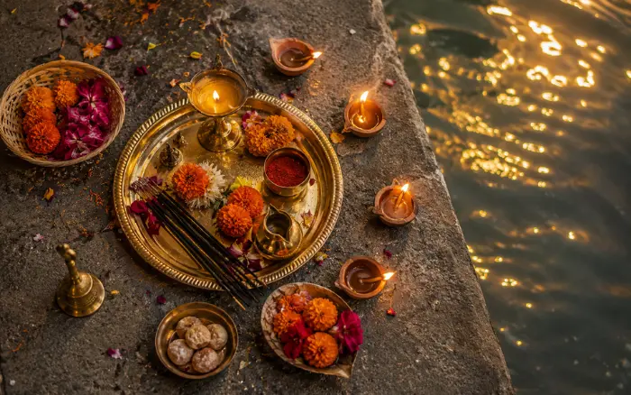 Traditional puja thali with brass lamp, marigold flowers, incense sticks and lit clay diyas placed on stone steps beside the Ganges River in Varanasi during Ganga Aarti